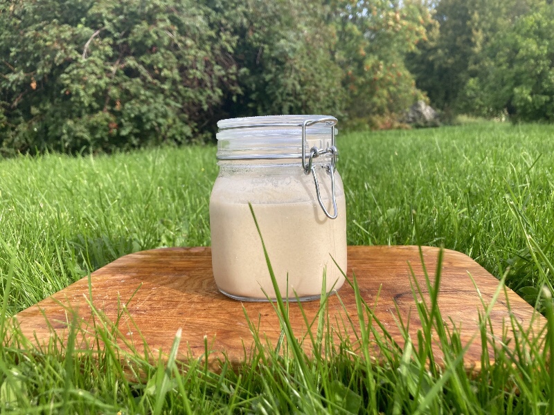 A jar of sourdough starter, looking like liquid dough, on top of a cutting board placed in green grass, against a backdrop of bushes and trees.